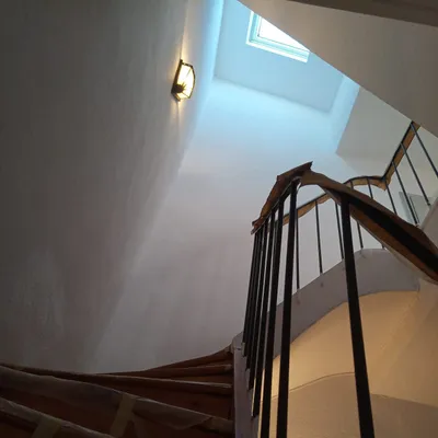 Cage d'escalier courbe vue d'en haut, murs blanc cassé, marches en bois protégées, une applique allumée