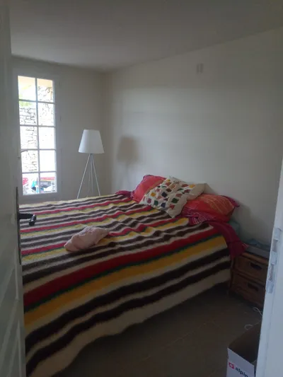 Chambre repeinte en blanc, lit avec couvre-lit rayé coloré, fenêtre donnant sur un muret en pierre