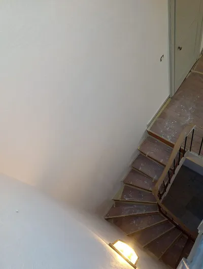 Escalier vu d'en haut, murs repeints en crème, marches en bois protégées, applique allumée en bas