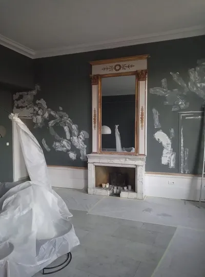 Salon en cours de peinture, mur vert sombre avec retouches, cheminée en marbre et miroir doré