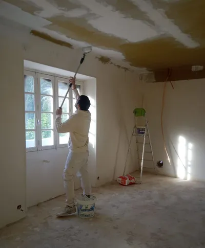 Peintre au rouleau sur perche traitant un plafond aux raccords d'enduit apparents, pièce en travaux