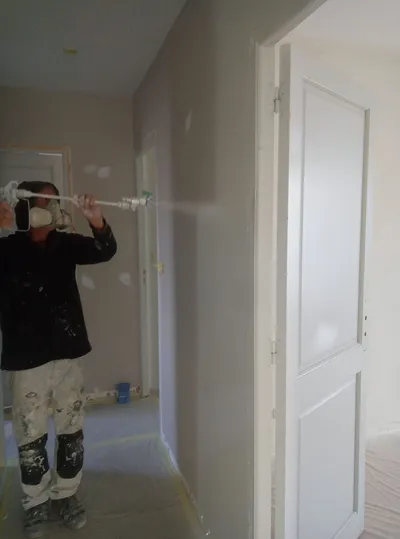 Peintre avec masque de protection pulvérisant un mur près d'une porte blanche, protections au sol