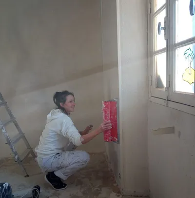 Artisane appliquant l'enduit à la taloche près d'une fenêtre, mur en cours de préparation