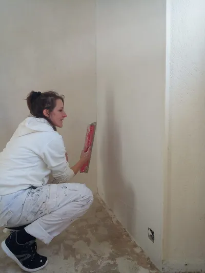 Artisane lissant l'enduit à la taloche dans l'angle d'une pièce aux murs beige clair