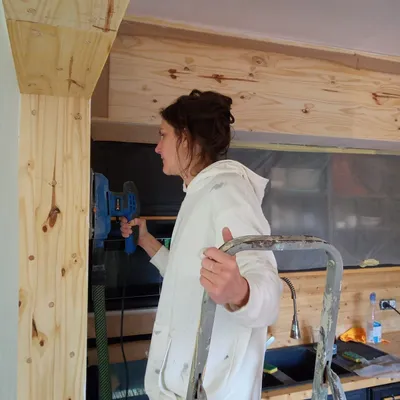 Artisane ponçant une poutre en bois sur un escabeau, préparation avant peinture