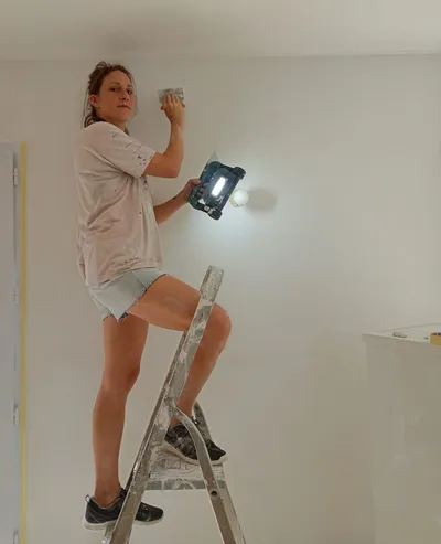 Artisane sur un escabeau, rebouchage d'un mur blanc à la spatule sous lampe d'appoint