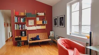 Salon vu en largeur, mur rouge peint avec étagères bois, fenêtre lumineuse et fauteuil corail