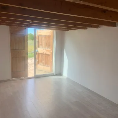 Pièce vide aux murs blancs fraîchement peints, plafond à poutres apparentes, porte ouverte sur le jardin