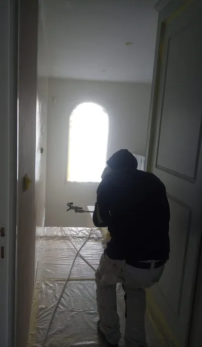 Peintre au pistolet dans une pièce protégée de bâches, silhouette devant une fenêtre en arc