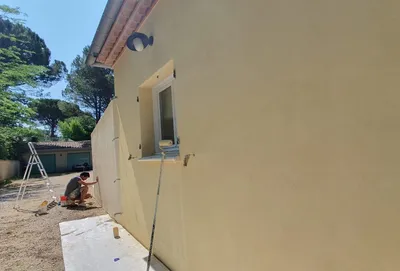 Façade extérieure couleur sable en cours de peinture, artisan agenouillé près du mur