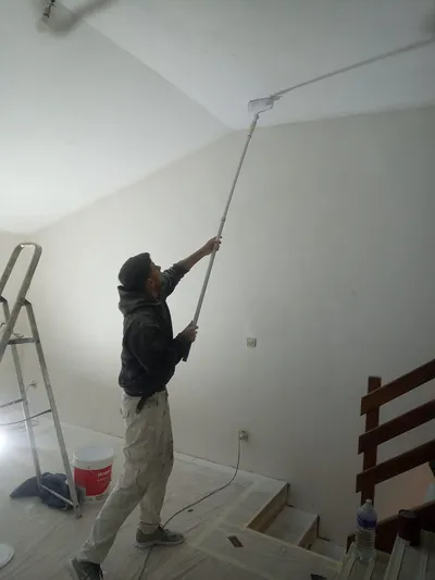 Peintre au rouleau à manche télescopique sur un plafond en pente, seau de peinture au sol